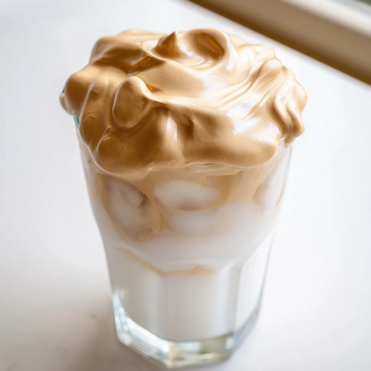 Fluffy whipped dalgona coffee floats atop cold milk, served in a glass with ice for a refreshing café-style treat.  