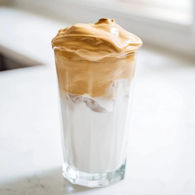 Whipped dalgona coffee with velvety peaks rests over milk, garnished with cocoa powder for a deliciously creamy, café-inspired sip.